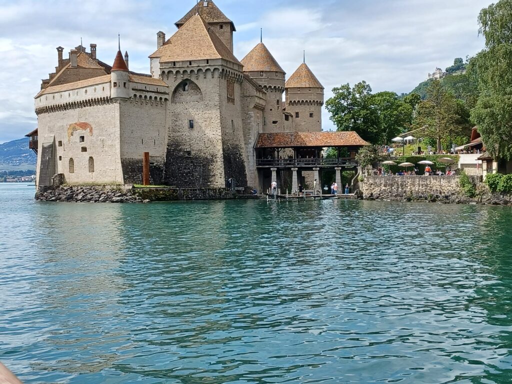Un château médiéval aux toits coniques se dresse au bord d'un lac, entouré d'eau claire et d'une verdure luxuriante sous un ciel nuageux.