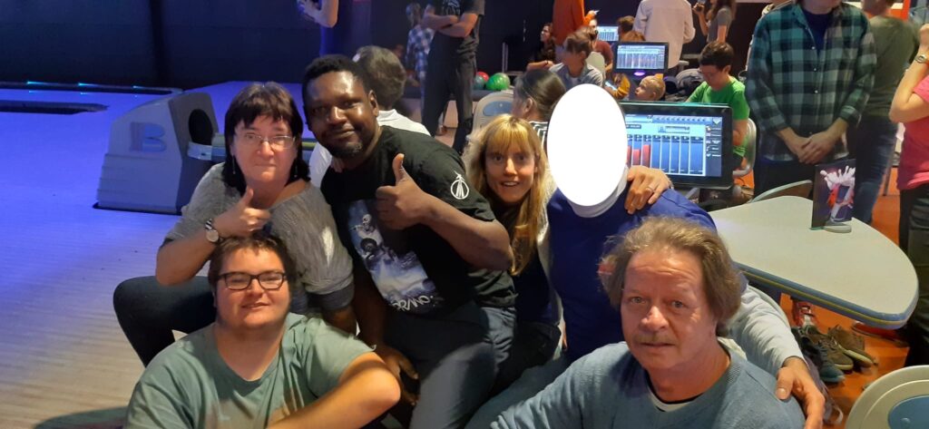 Groupe de personnes rassemblées dans un bowling, certaines assises et d'autres debout, avec des pistes de bowling et des écrans visibles en arrière-plan.