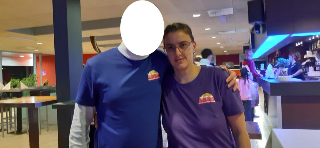 Deux personnes portant des t-shirts bleus et violets se tiennent côte à côte dans un bar. Le visage d'une personne est caché. Le bar est éclairé par un éclairage bleu.