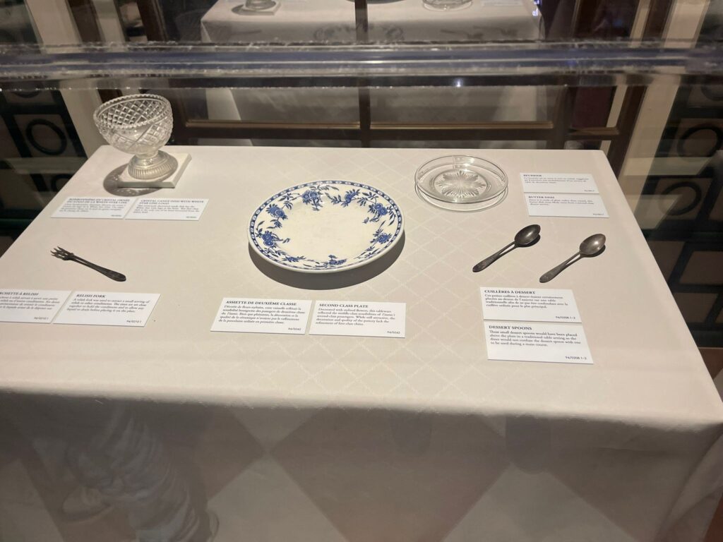 Vitrine présentant de la vaisselle historique, comprenant une assiette à motifs, deux cuillères, une fourchette, un plat en verre et un petit bol, chacun avec des étiquettes descriptives.