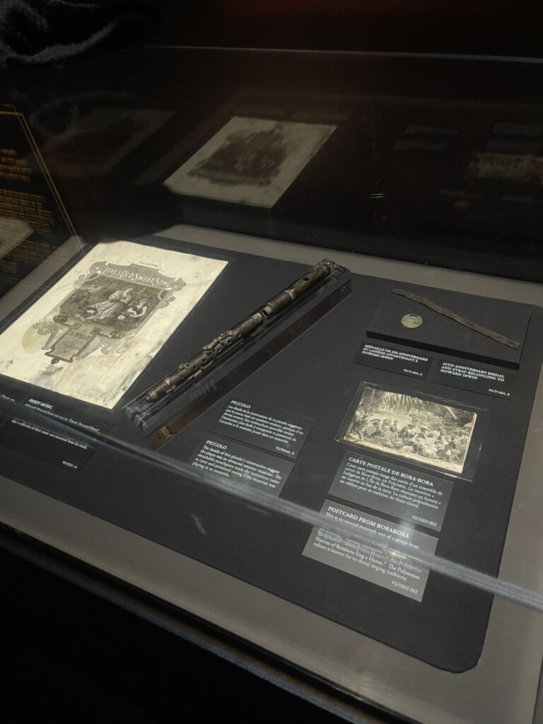 Vitrine contenant des documents historiques, un coffret en bois, une pièce de monnaie et un petit objet noir avec des étiquettes informatives sur fond sombre.