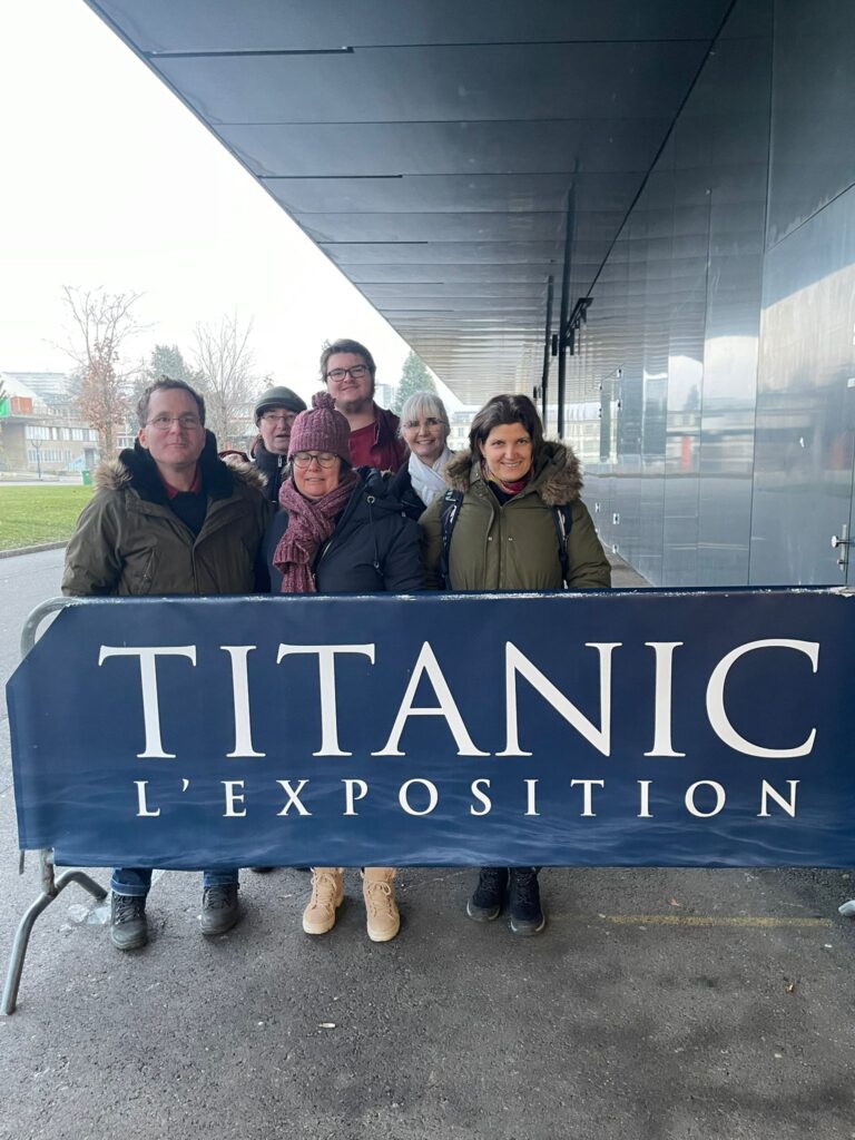 Un groupe de personnes debout derrière un panneau indiquant « Titanic L'Exposition » à l'extérieur d'un bâtiment moderne.